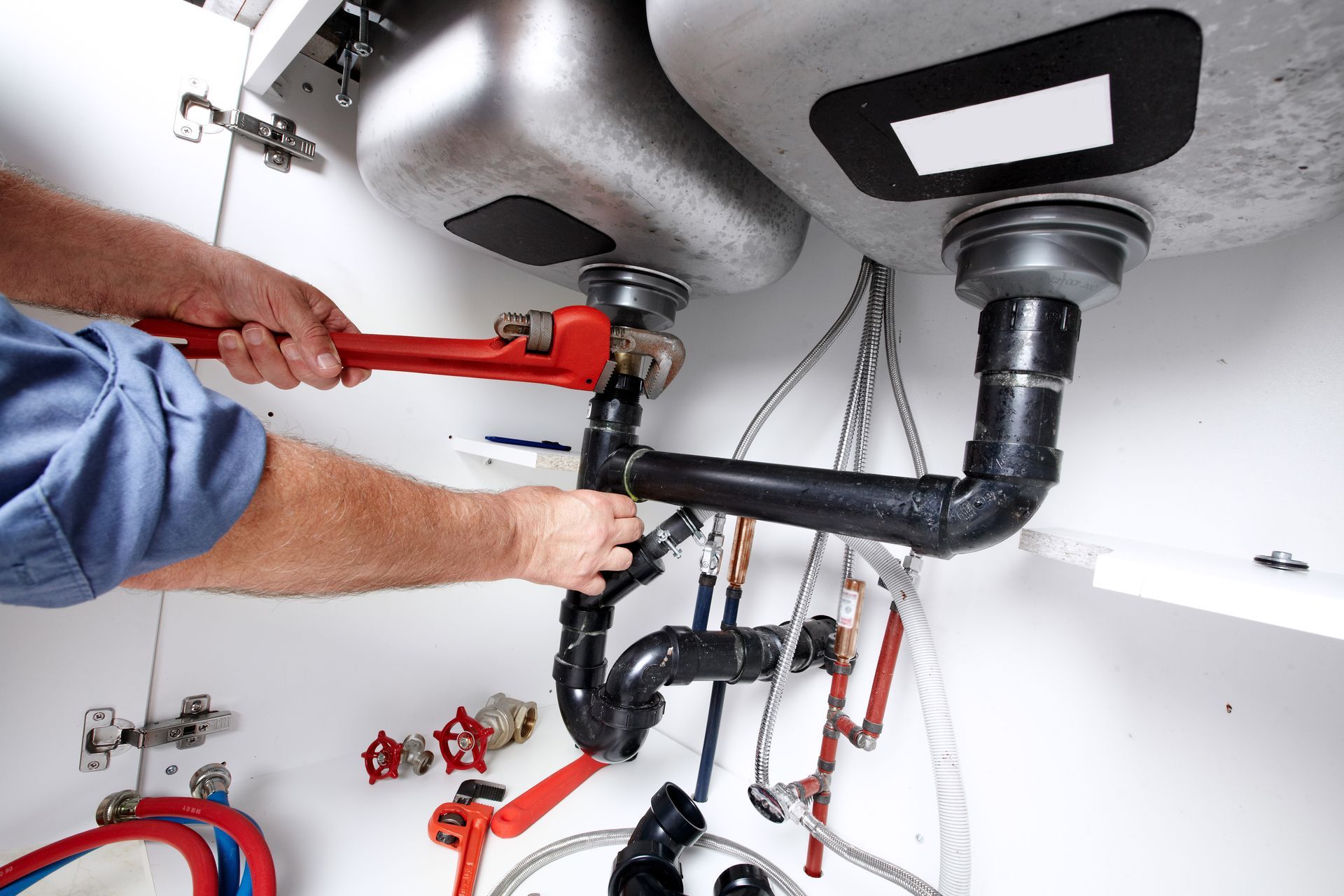 A plumber tightening black drain and supply pipes under a double sink using a red wrench