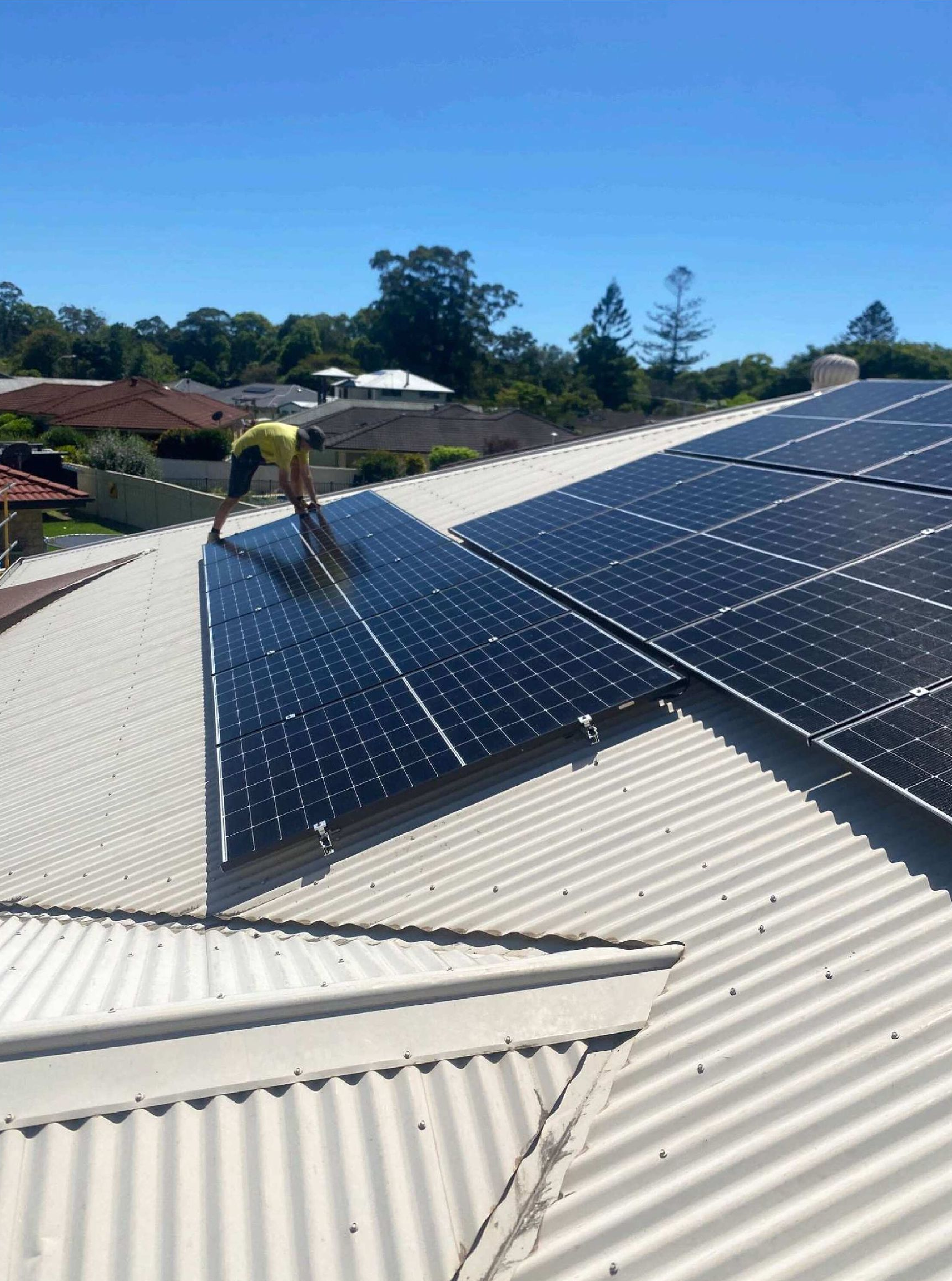 Person installing solar panels on a corrugated metal roof under a blue sky— Ray Smith Electrical in Korora, NSW