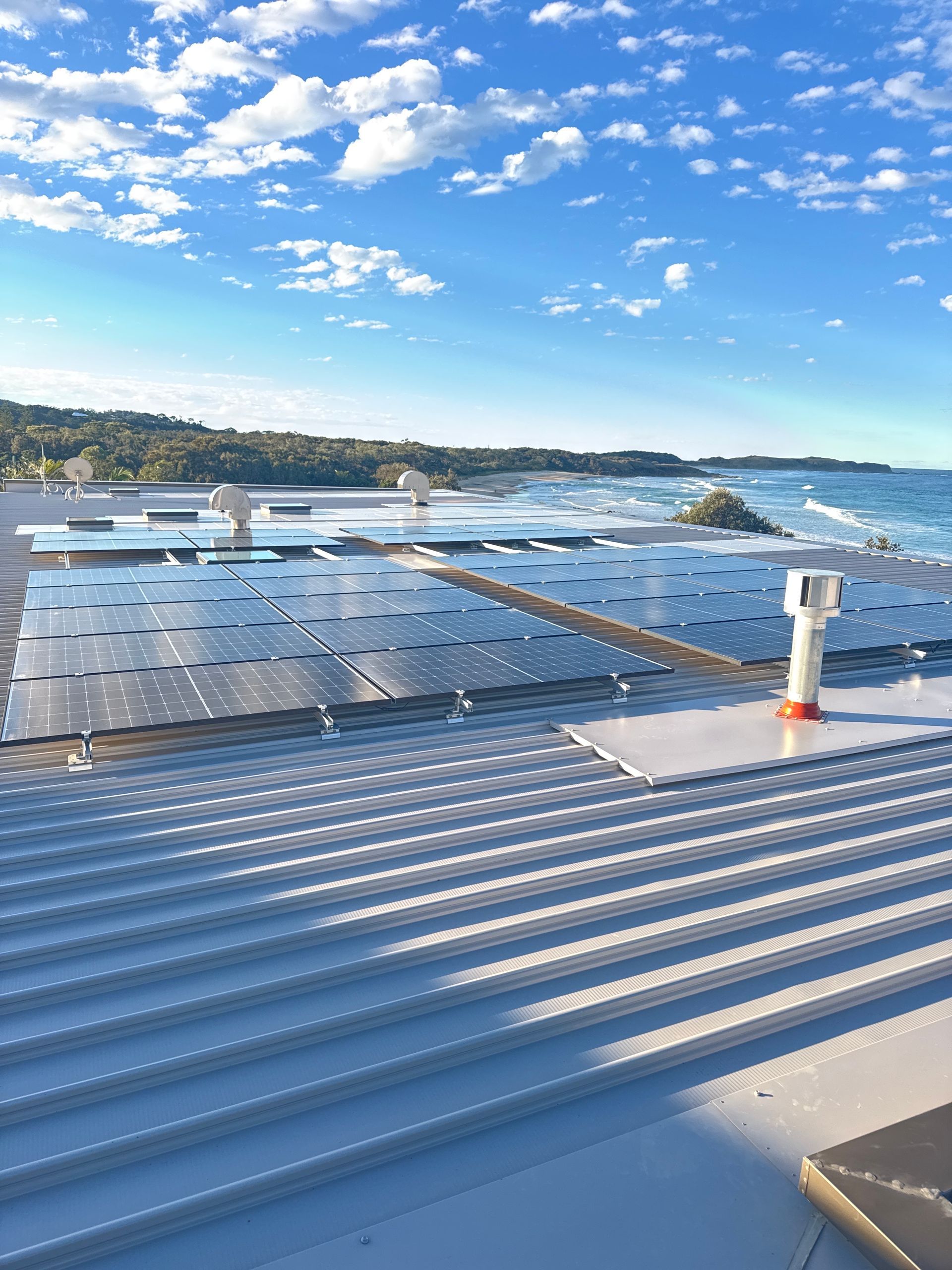 Solar panels on a building rooftop overlooking a blue ocean under a bright, cloudy sky— Ray Smith Electrical in Korora, NSW