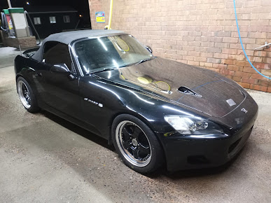 Black Honda S2000 convertible with carbon fiber hood parked at a car wash, brick wall in background.