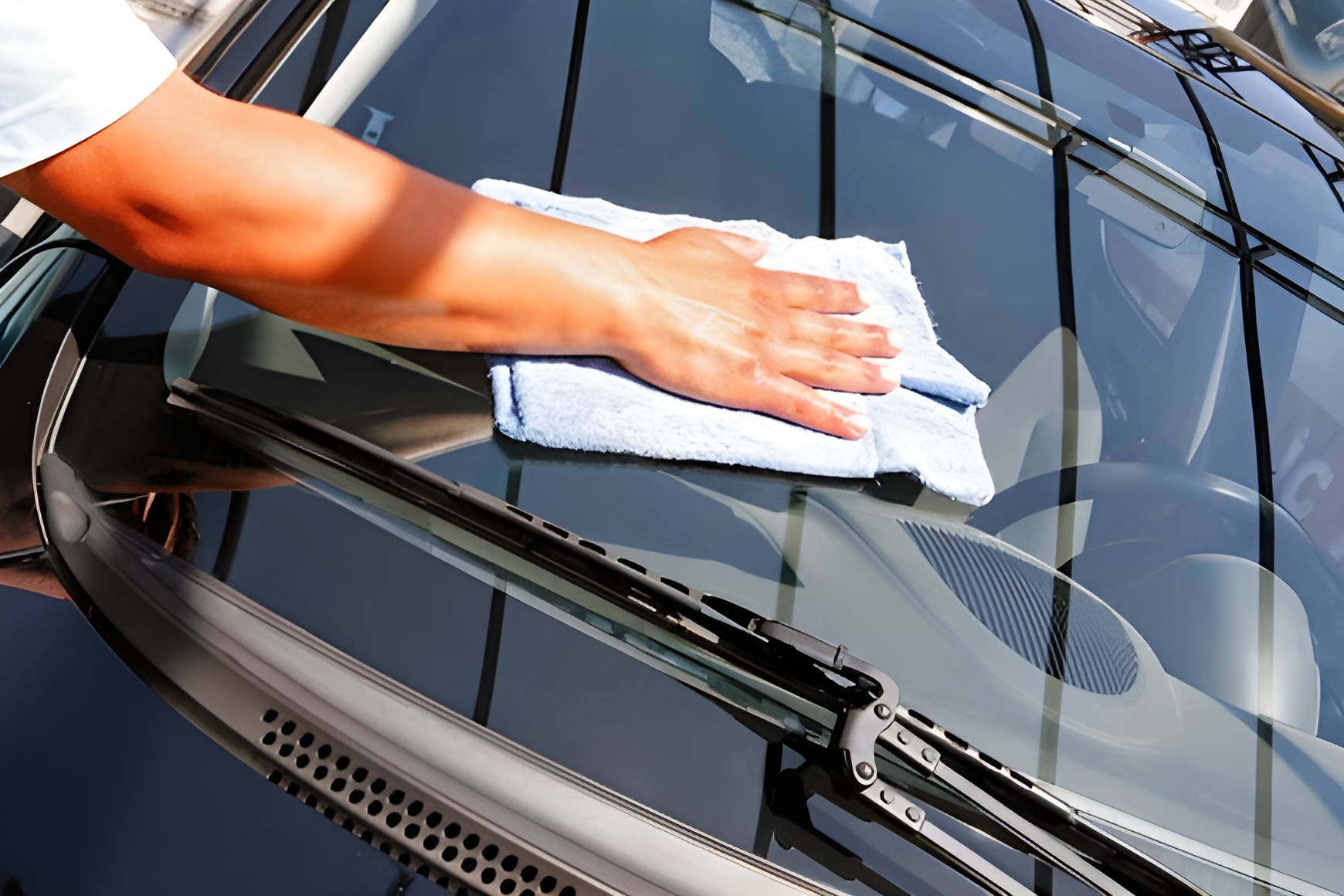 Person wiping a car's windshield with a blue cloth. Sunlight reflects off the dark car.