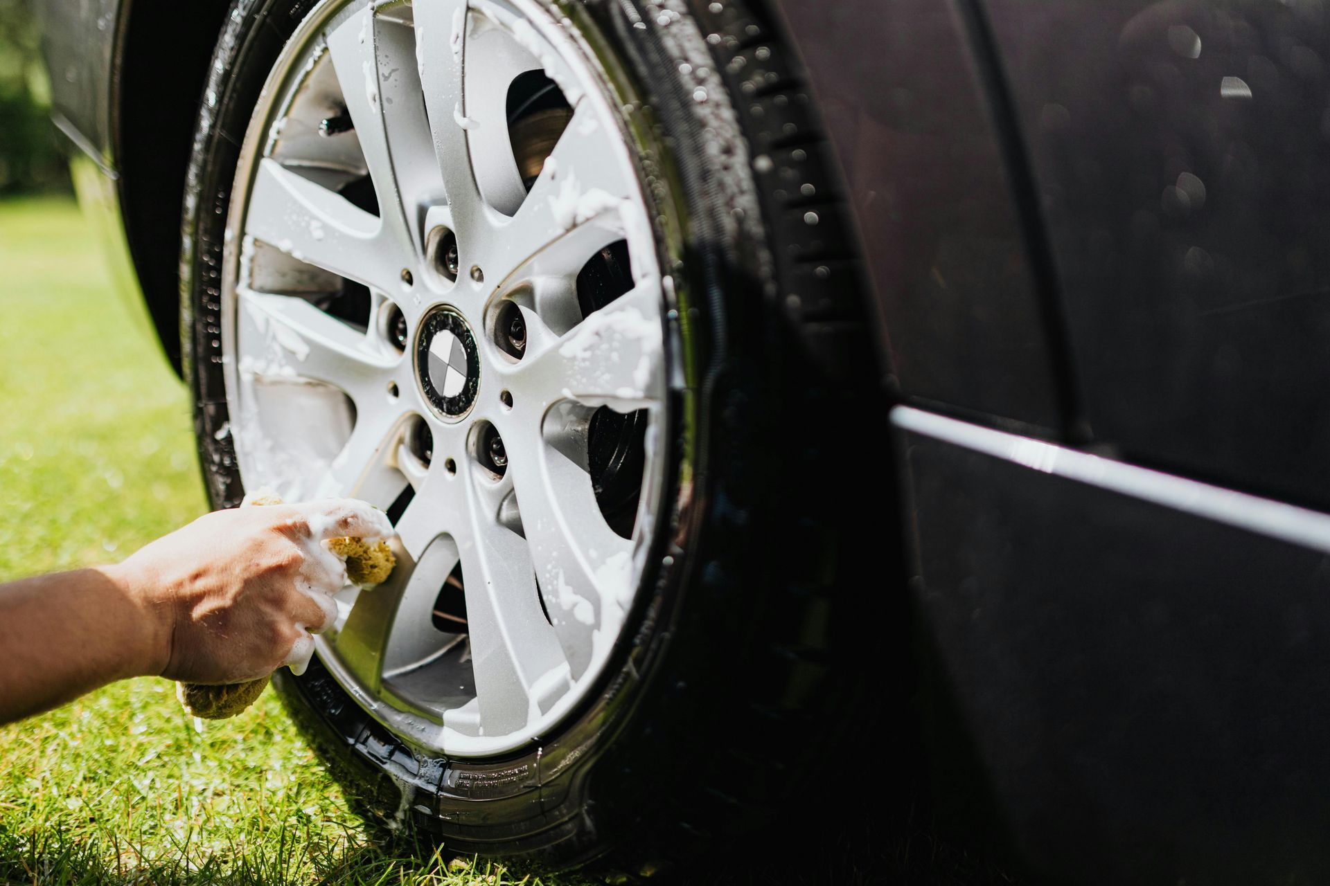 Person washing a car's tire with a soapy sponge outdoors on a grassy lawn.