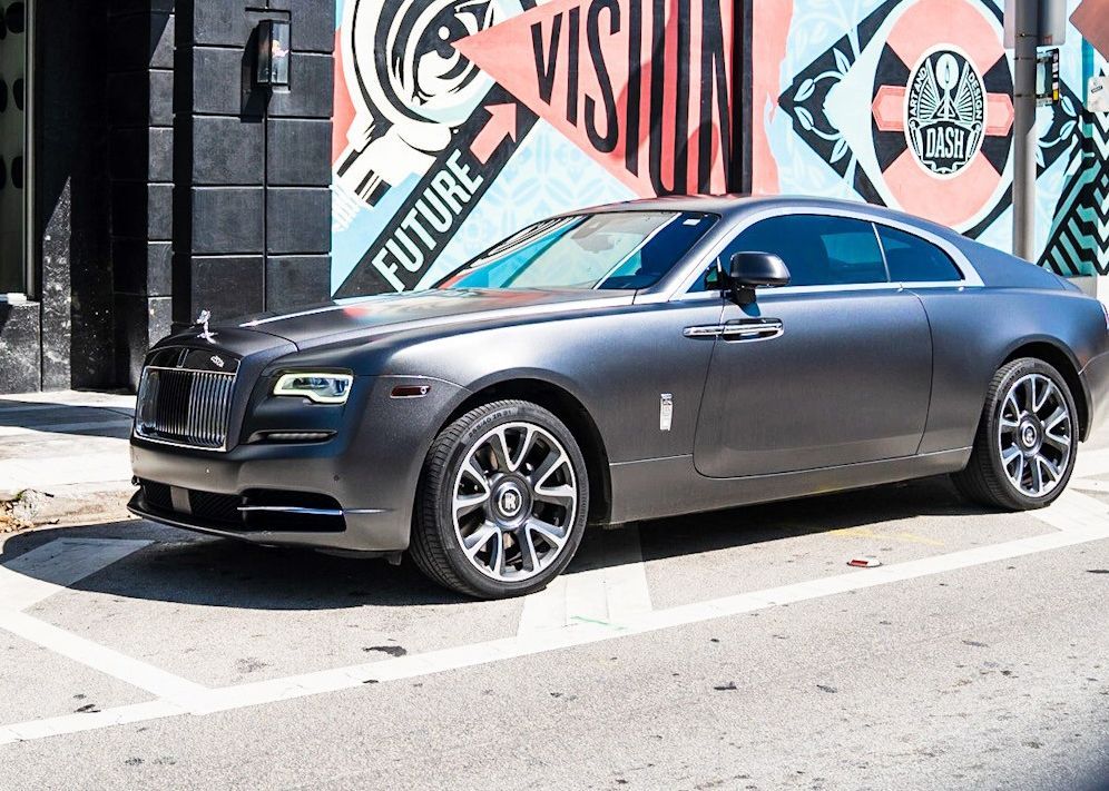 A gray Rolls Royce Wraith is parked in front of a building with a mural on it.
