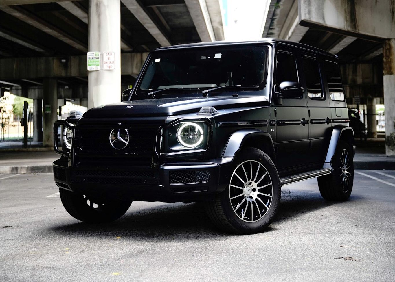 A black Mercedes Benz G Class is parked under a bridge in a parking lot.