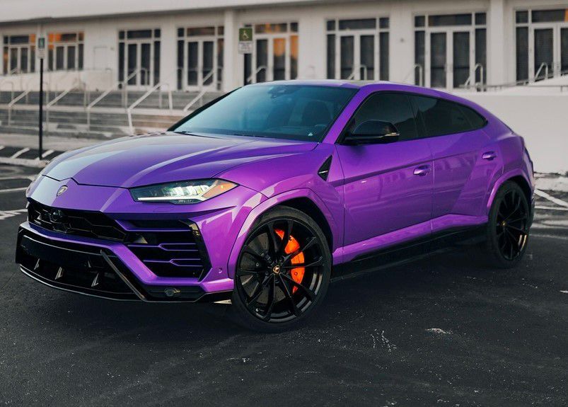 A purple Lamborghini Urus is parked in a parking lot in front of a building.