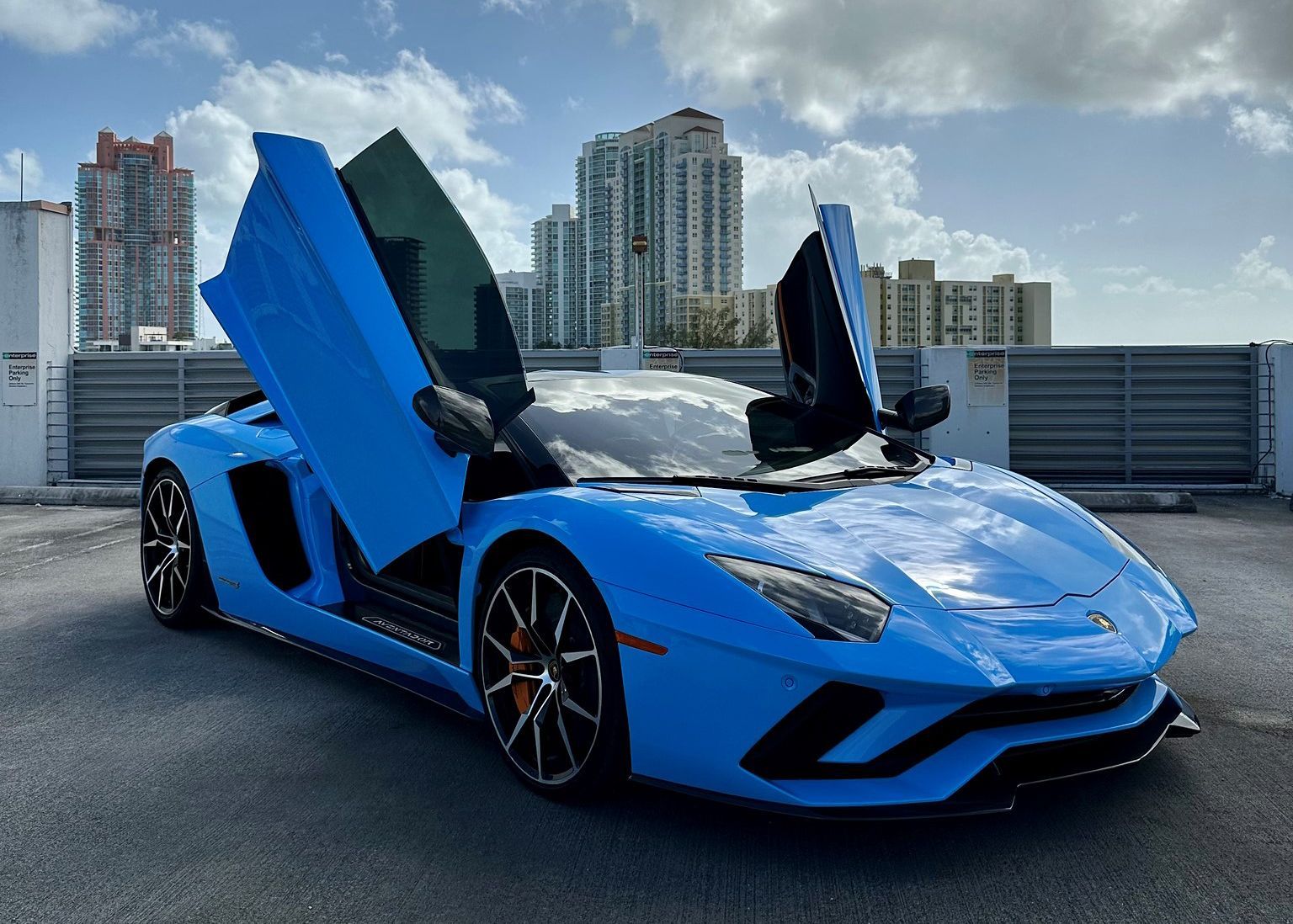A blue Lamborghini Aventador with its doors open is parked in a parking lot.