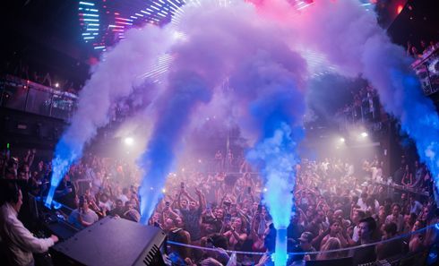 A crowd of people at a concert with smoke coming out of the stage.