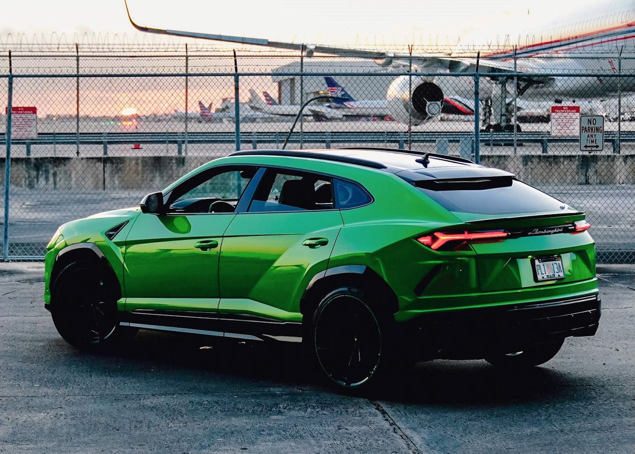 A green Lamborghini Urus is parked in a parking lot with an airplane in the background.