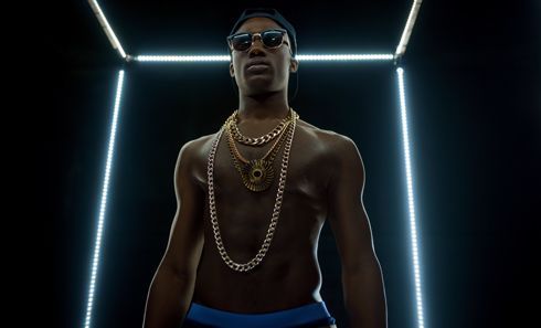 A shirtless man wearing sunglasses and a gold chain is standing in a dark room.