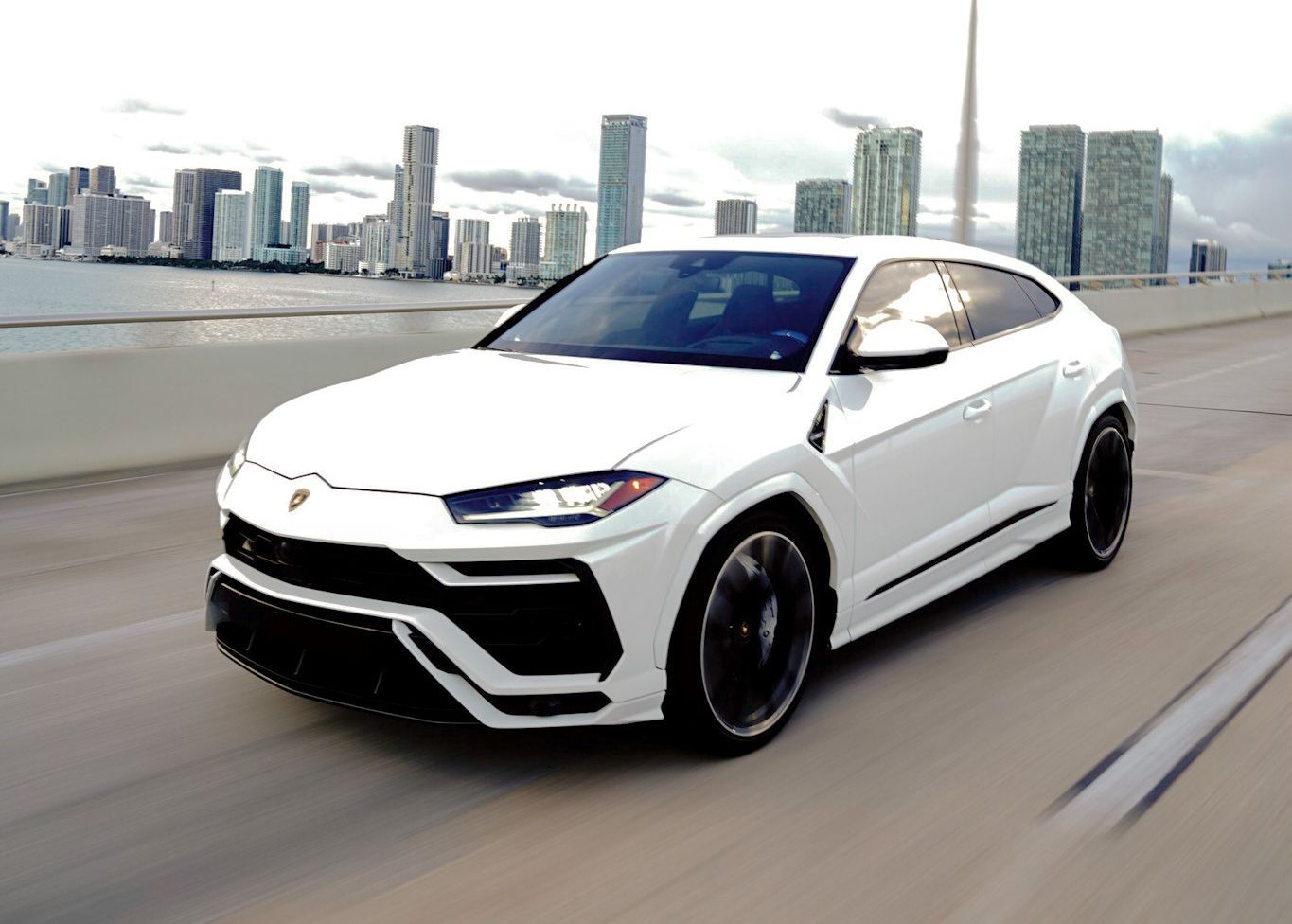 A white Lamborghini Urus is driving on a highway in front of a city skyline.