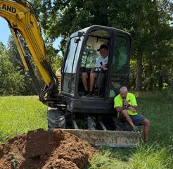 Two people take a break near a yellow excavator in a grassy, outdoor area next to a pile of excavated dirt.