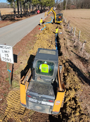 A yellow skid steer and excavator work to install a blue water pipe in a dirt trench beside a rural road.