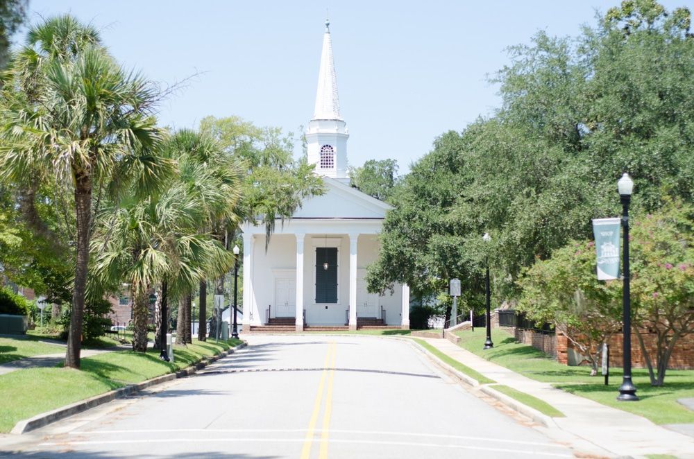 White church with steeple at end of road lined with trees under a bright sky.