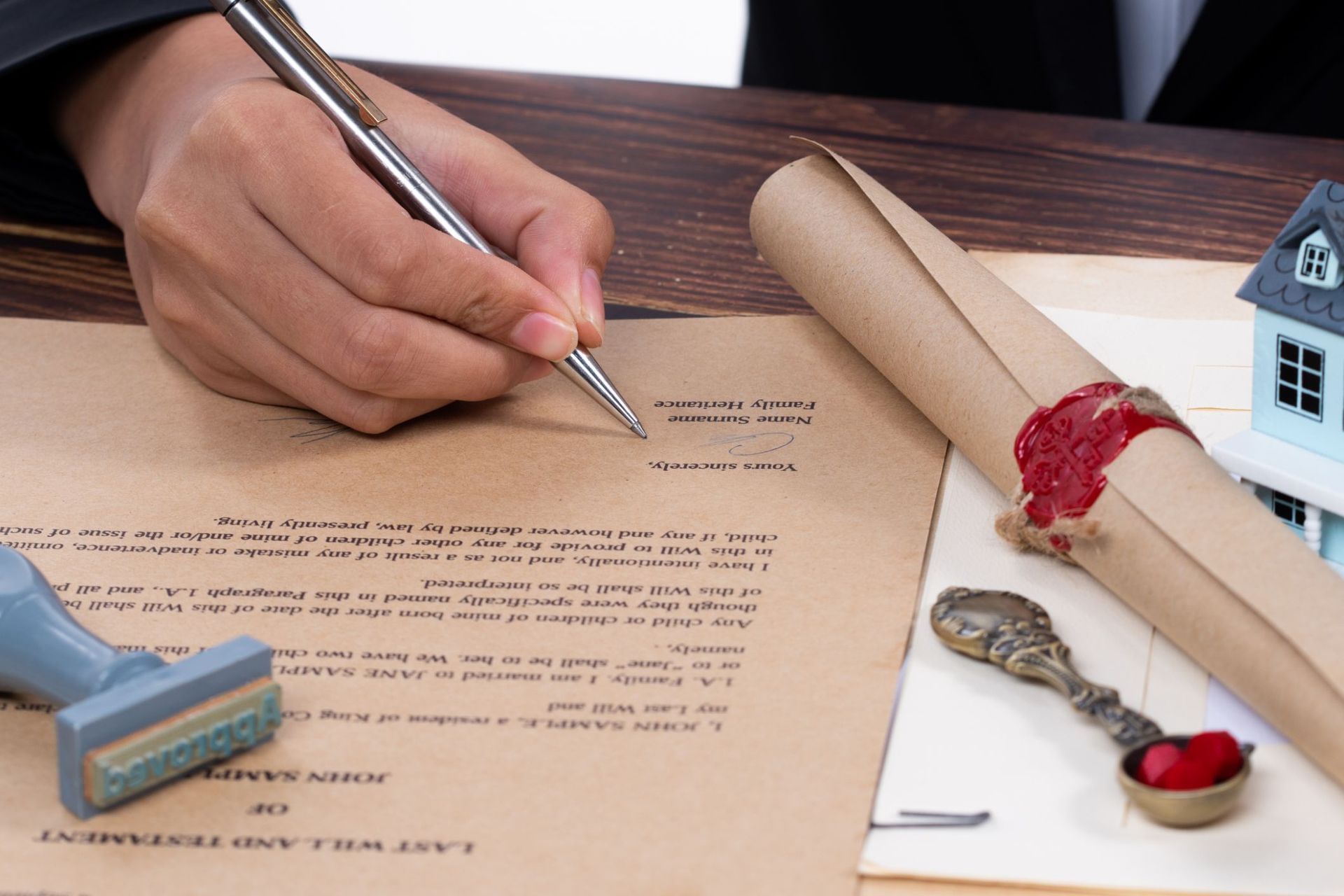 Person signing a document with pen, next to a sealed scroll, stamp, and miniature house on wooden surface.