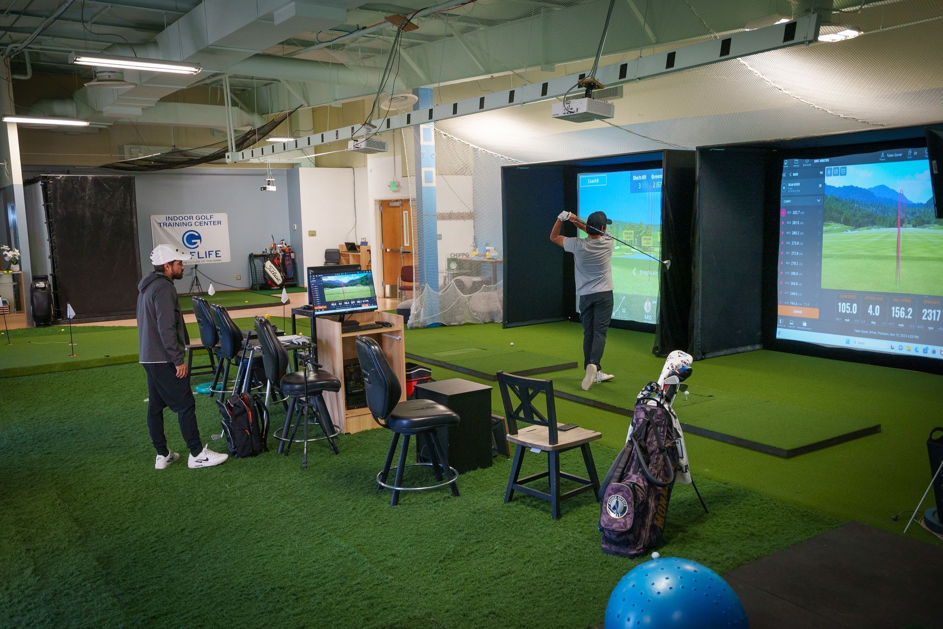 Indoor golf training facility, man swinging club at a screen, others watch. Artificial turf, chairs, and golf bags.