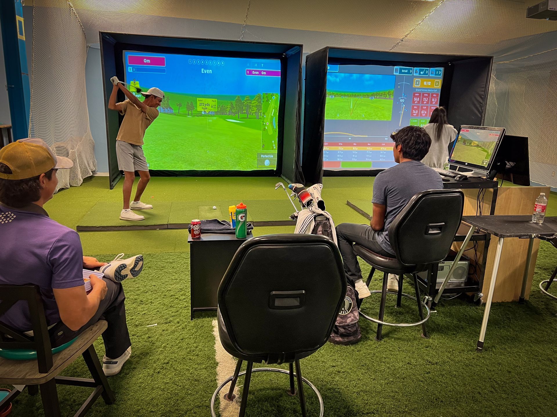 Golfers in an indoor simulator, hitting and watching screens. Others observe from chairs. Green turf.