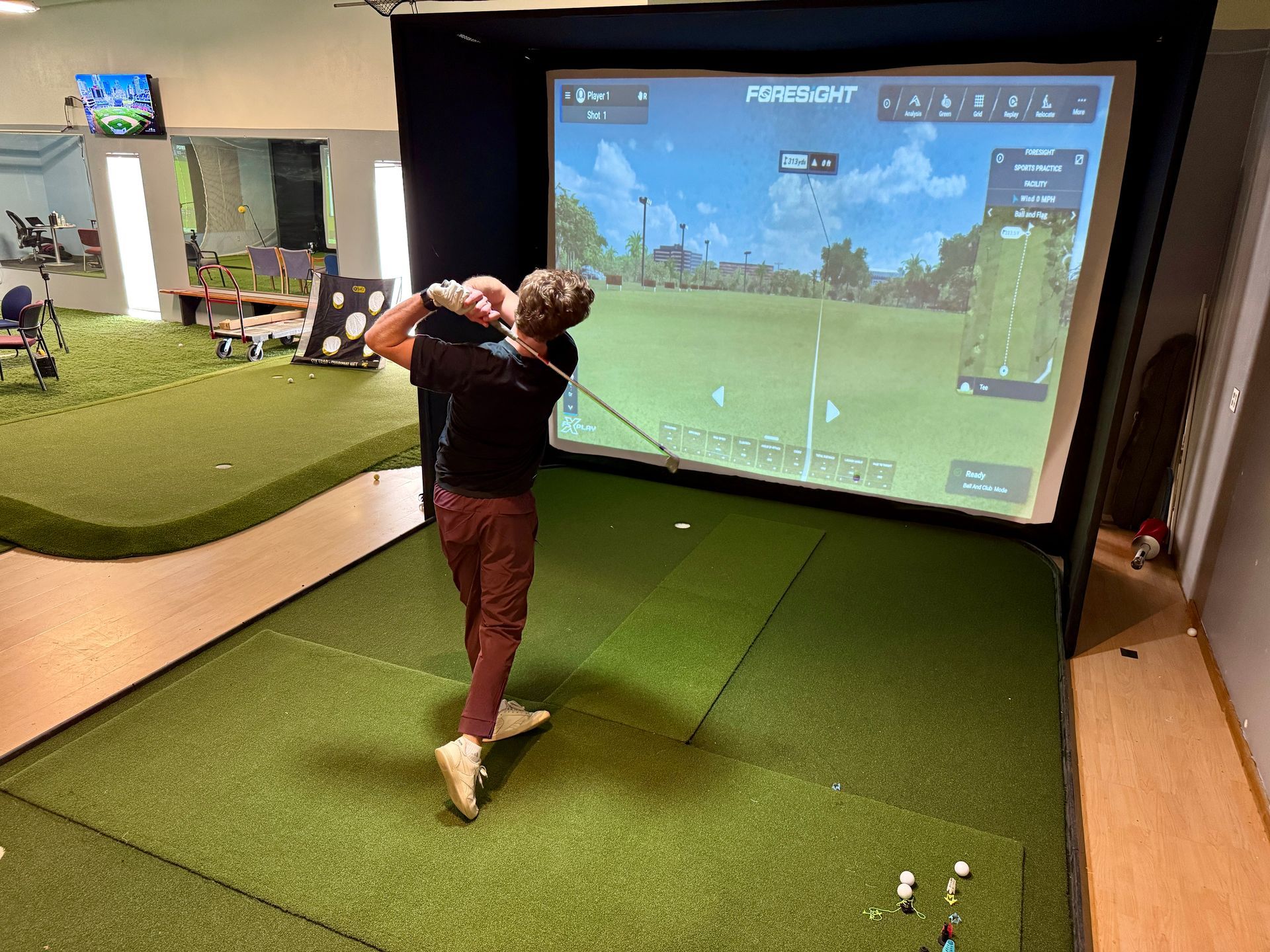 A man swings a golf club in an indoor golf simulator, hitting towards a screen displaying a course.