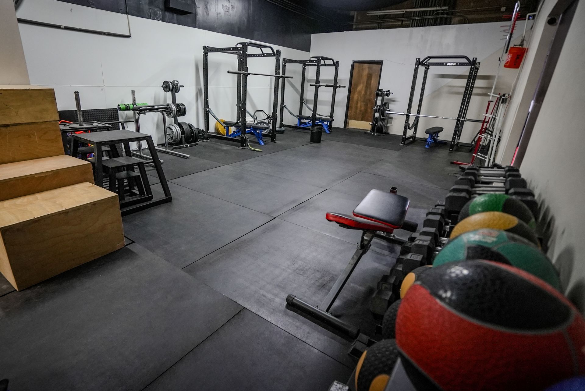 A gym with black rubber flooring, workout racks, medicine balls, and wooden boxes.