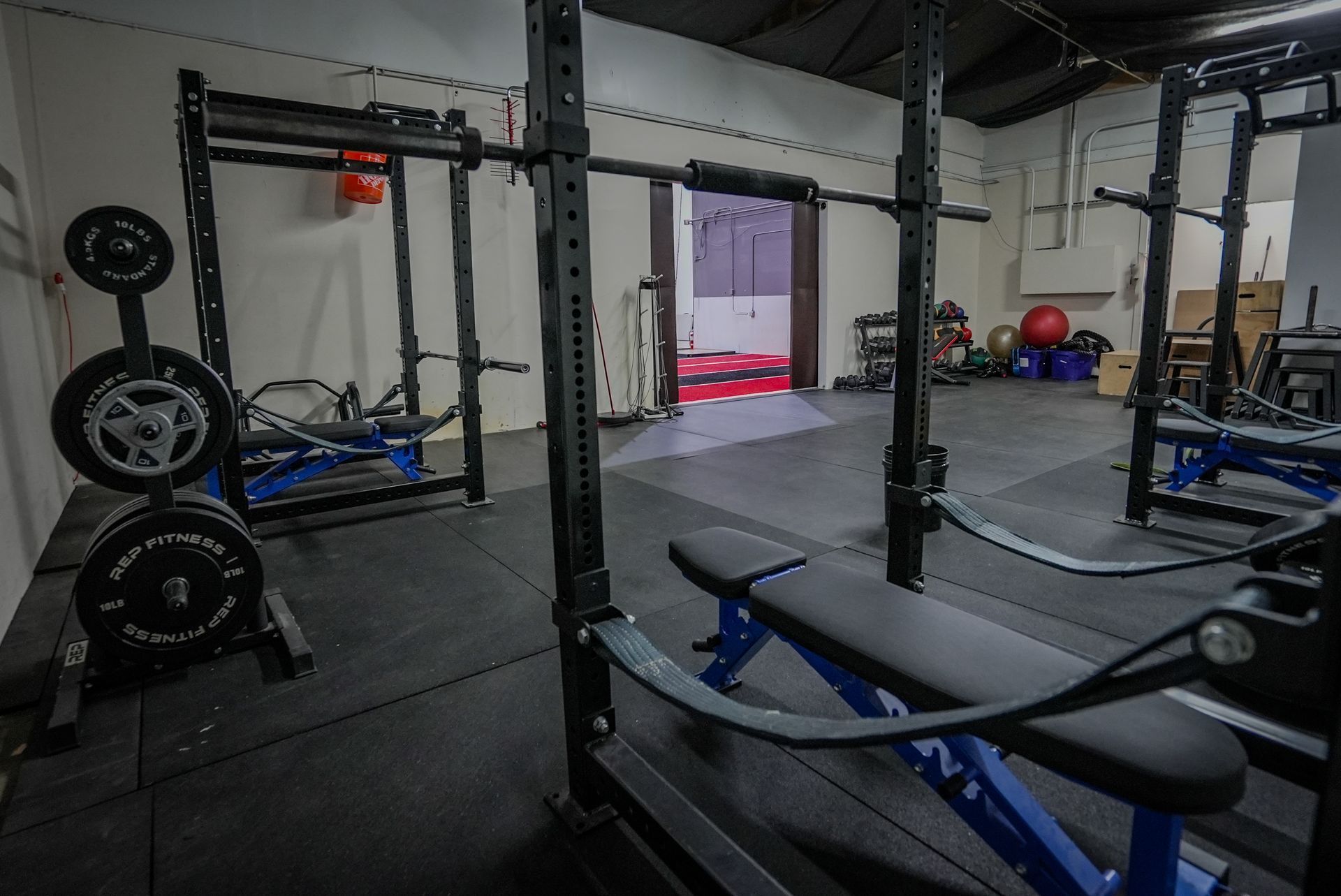 Weightlifting gym with black squat racks, blue benches, weights, and rubber flooring.