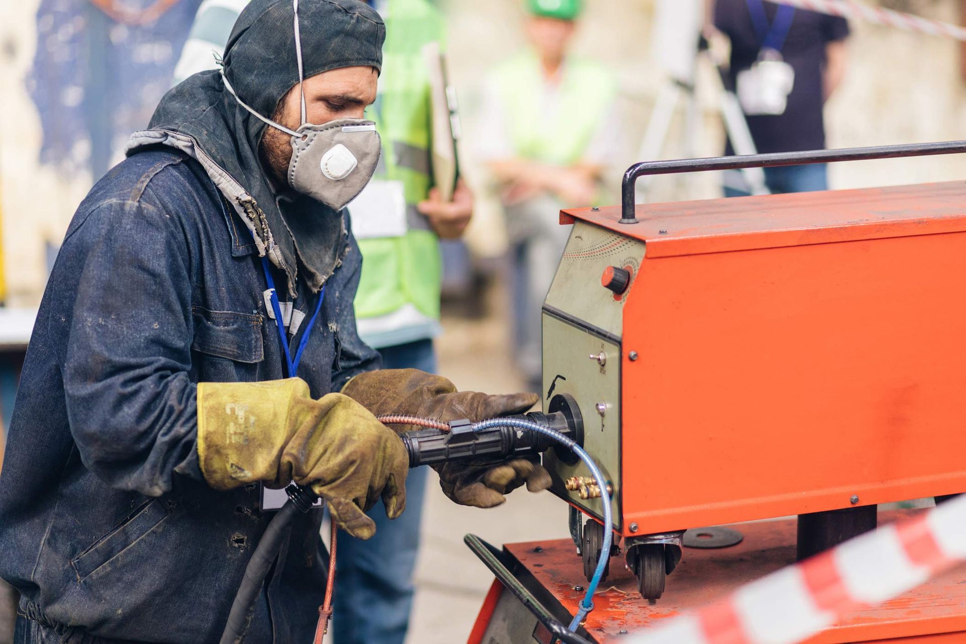 Welder wearing safety gear uses an orange machine outdoors, other people in background.