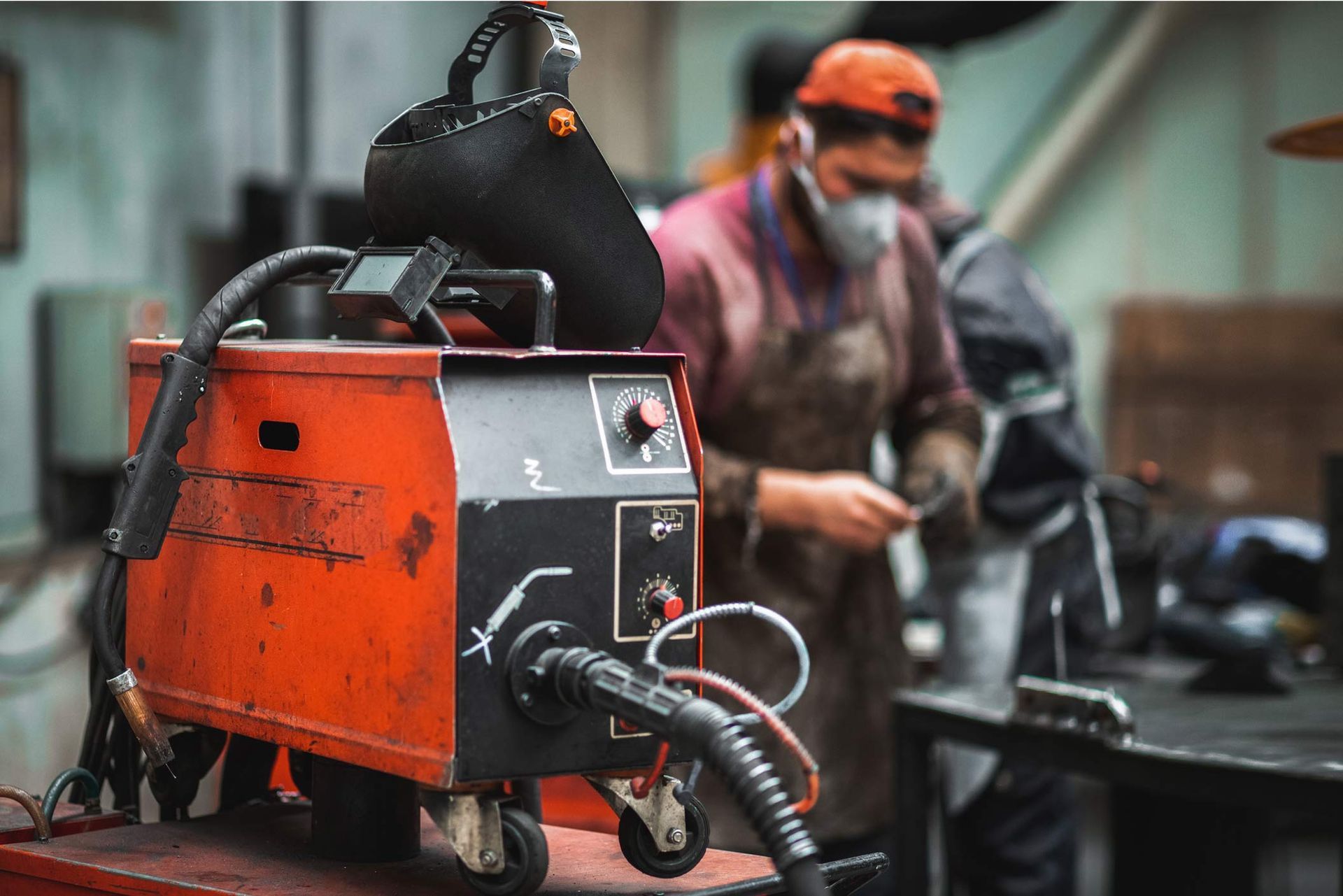 Red welding machine in a workshop, with a worker in the background wearing a mask.