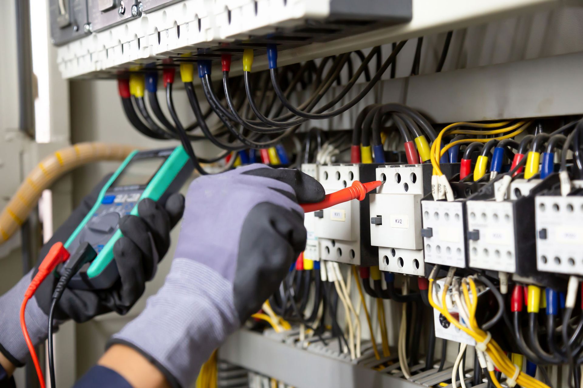Electrician tests wiring in a control panel, wearing gloves and using a multimeter.