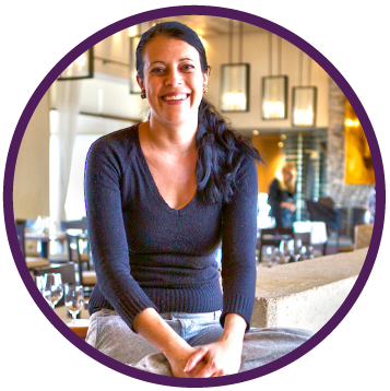 Woman with dark hair smiles at a restaurant, sitting on a ledge. Purple circle frame.