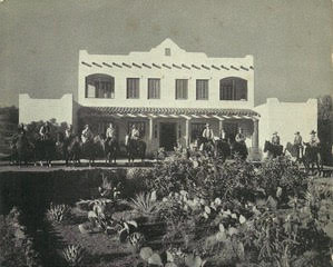 A black and white photo of people riding horses in front of a building.