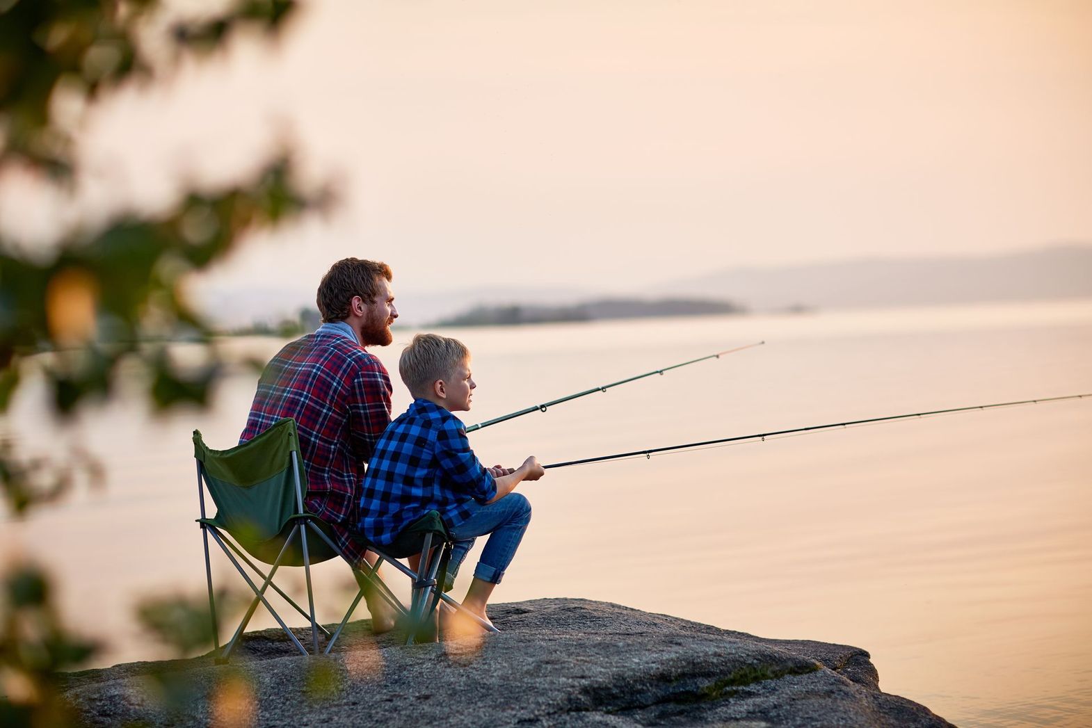 Two people sit on a rock, fishing together at sunset by a calm lake.