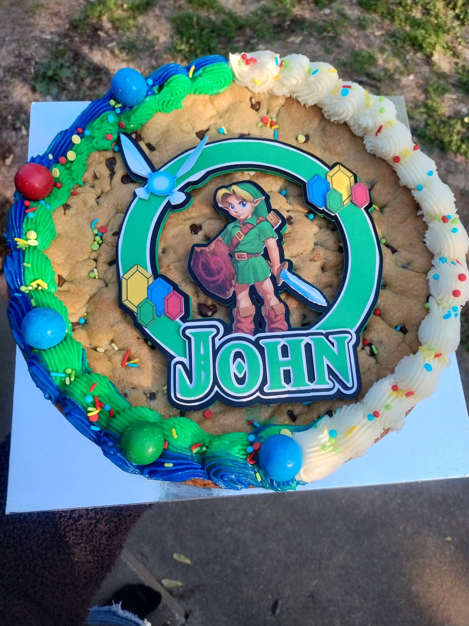 Giant Link-themed cookie cake with "John" on it, decorated with blue, green, and white frosting and candy.
