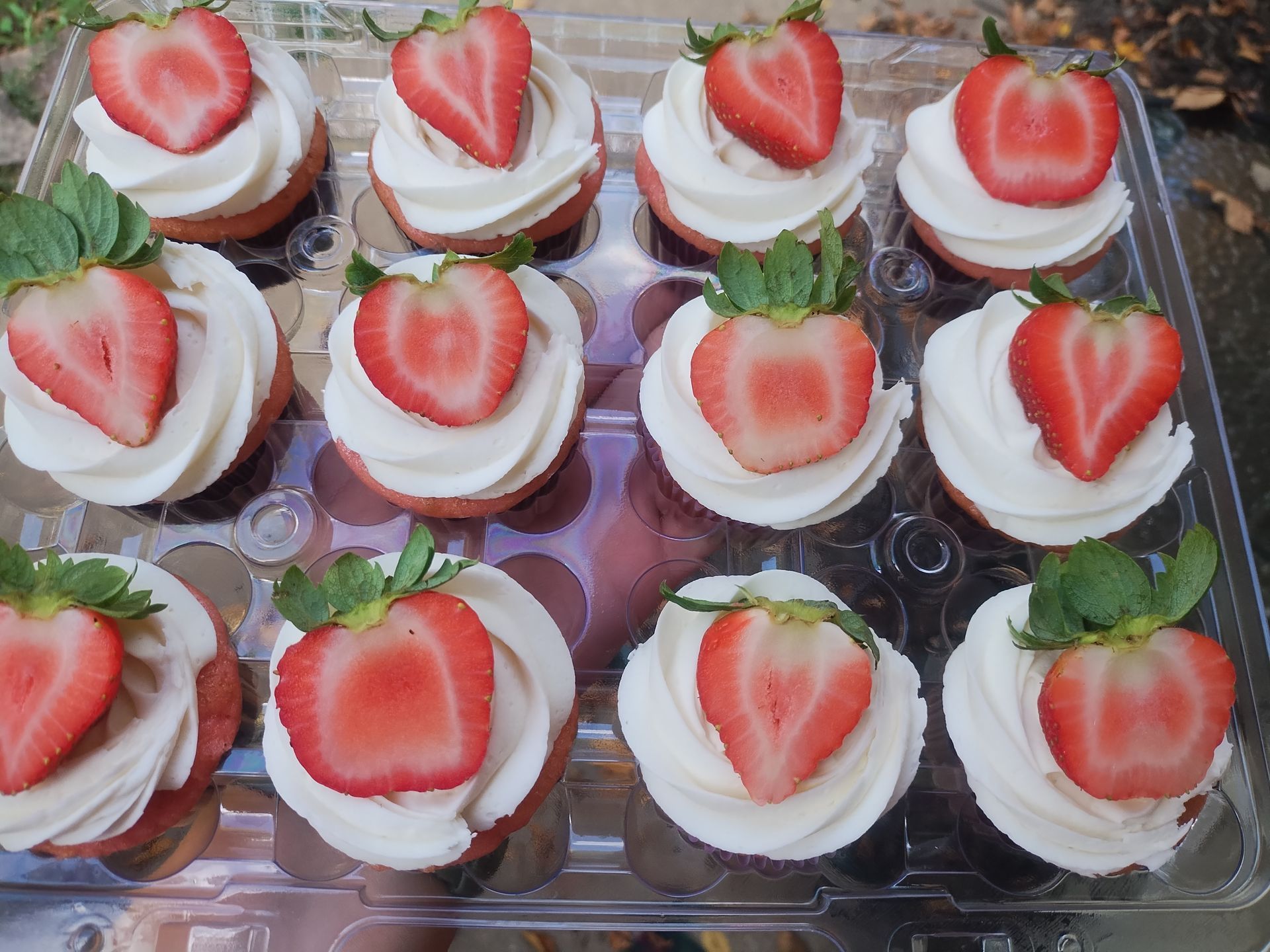 Strawberry cupcakes in a clear plastic container, topped with white frosting and strawberry halves.