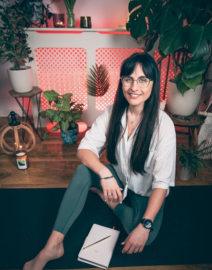 A woman is sitting on the floor in front of a fireplace surrounded by potted plants.