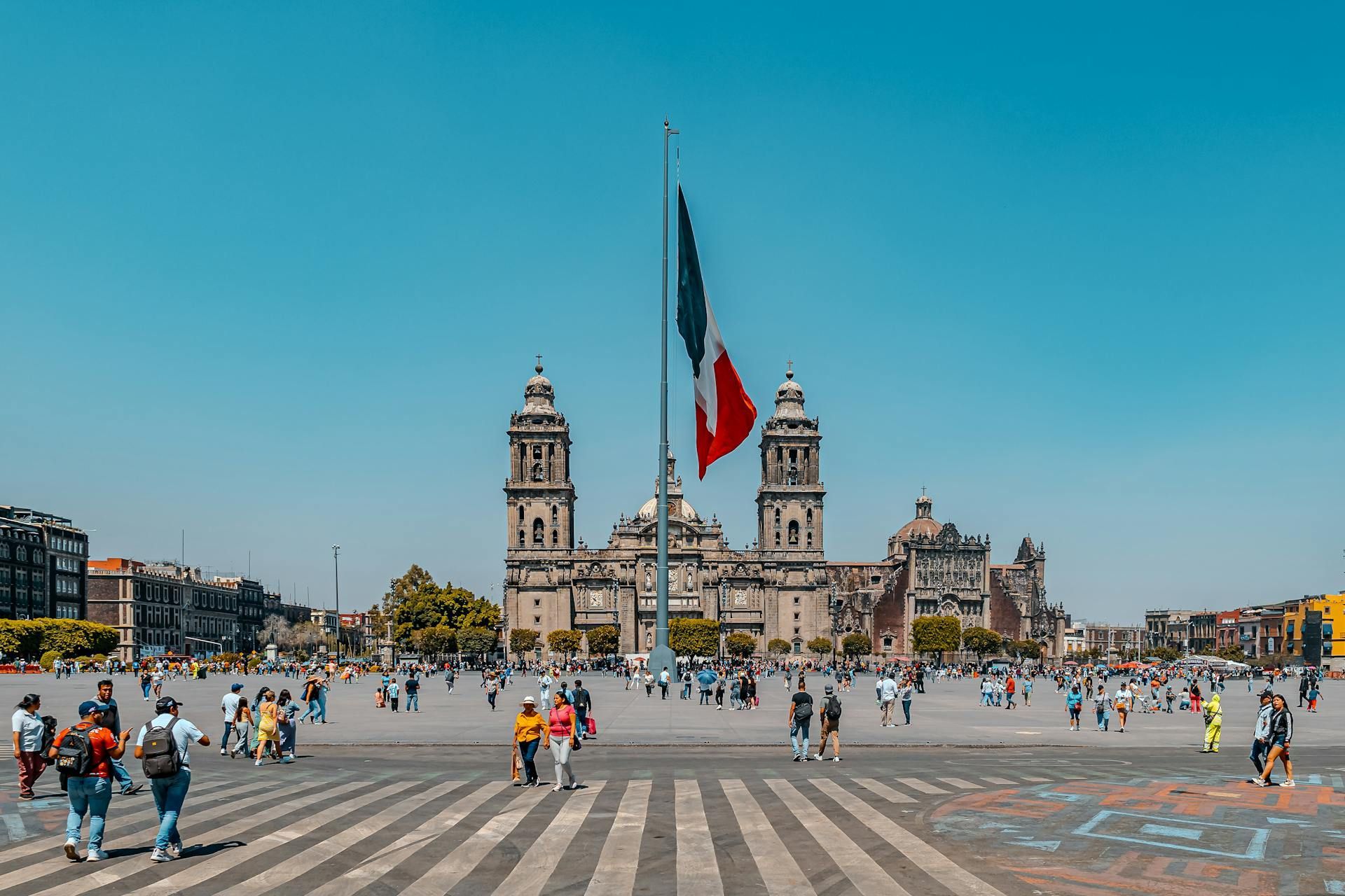 Una bandera mexicana ondea frente a un gran edificio en una plaza de la ciudad.