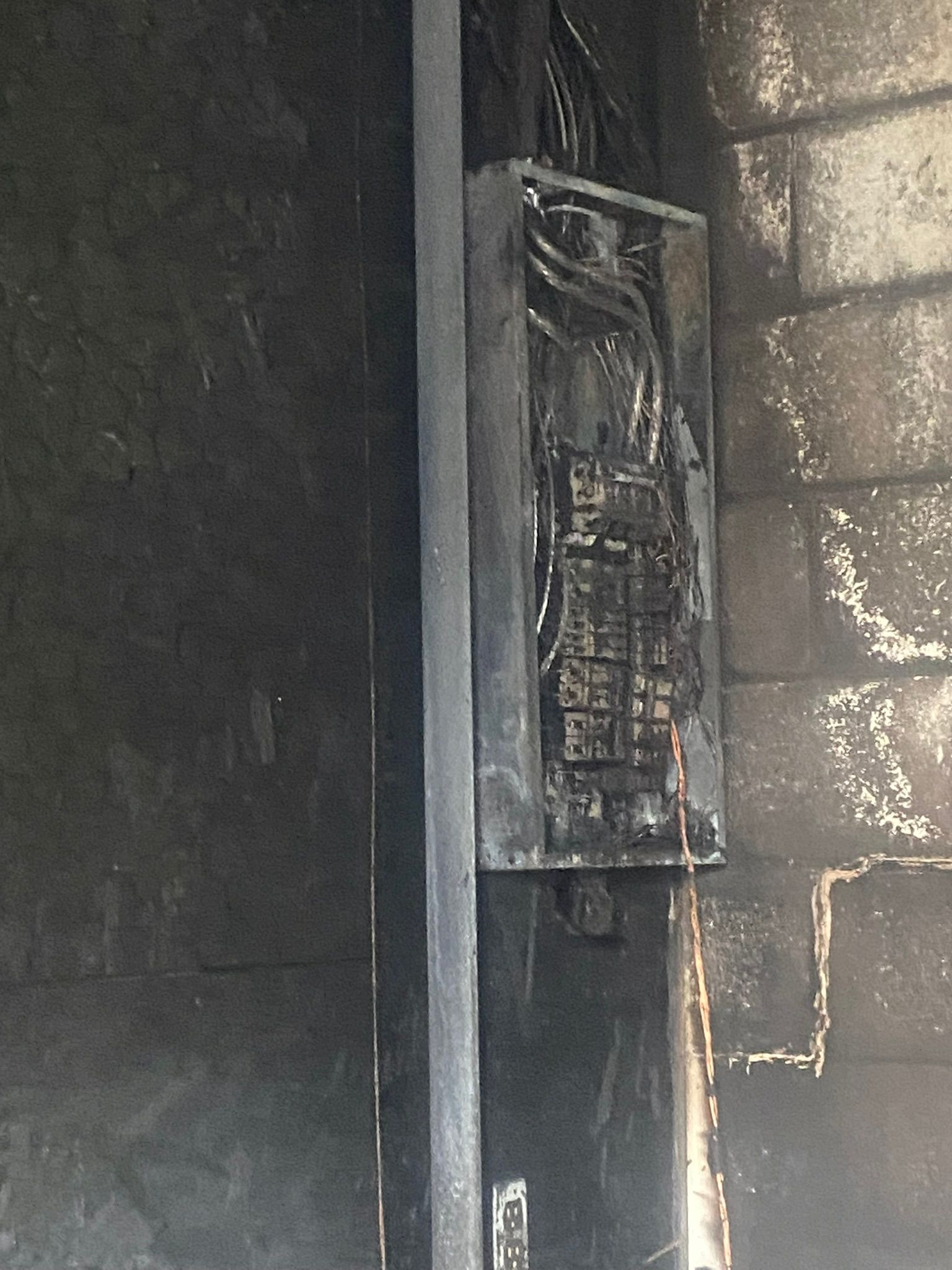 A broken electrical box is hanging on a brick wall.