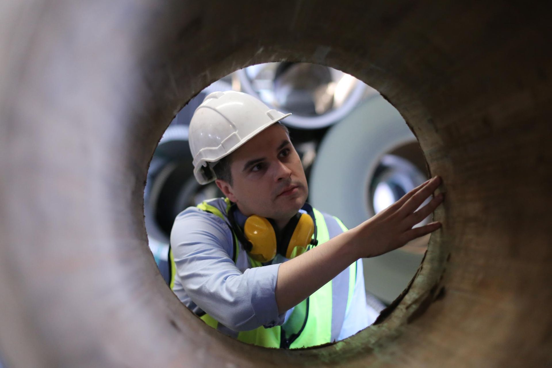A man wearing a hard hat and ear muffs is looking through a hole in a pipe.