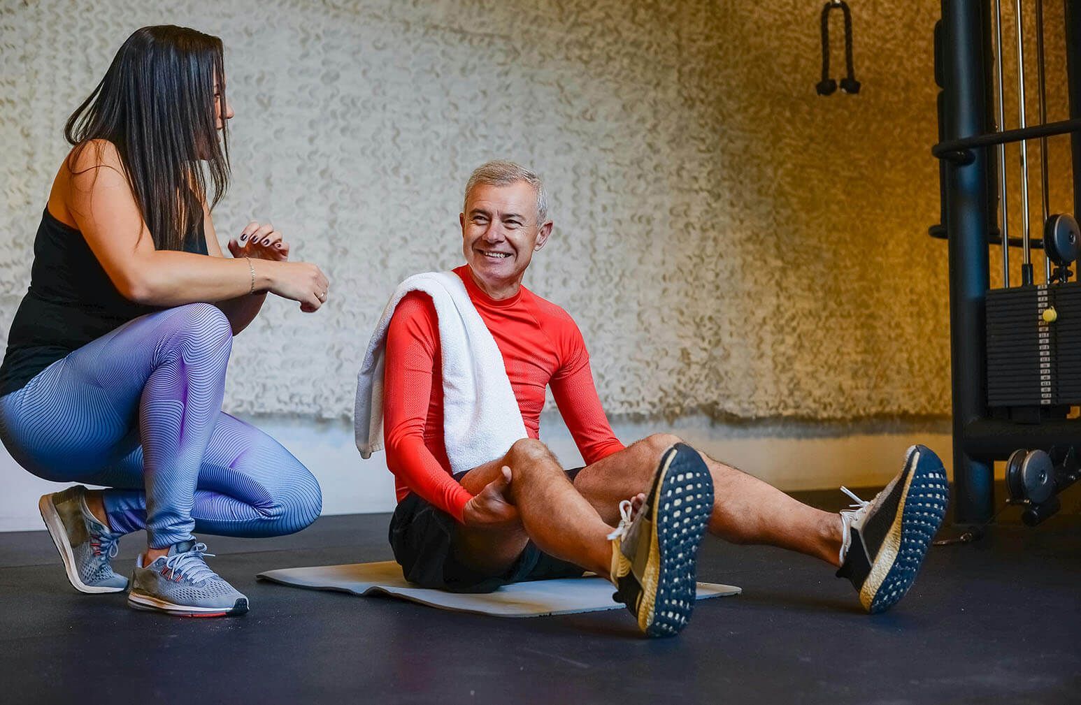 A woman is squatting next to a man sitting on the floor in a gym.