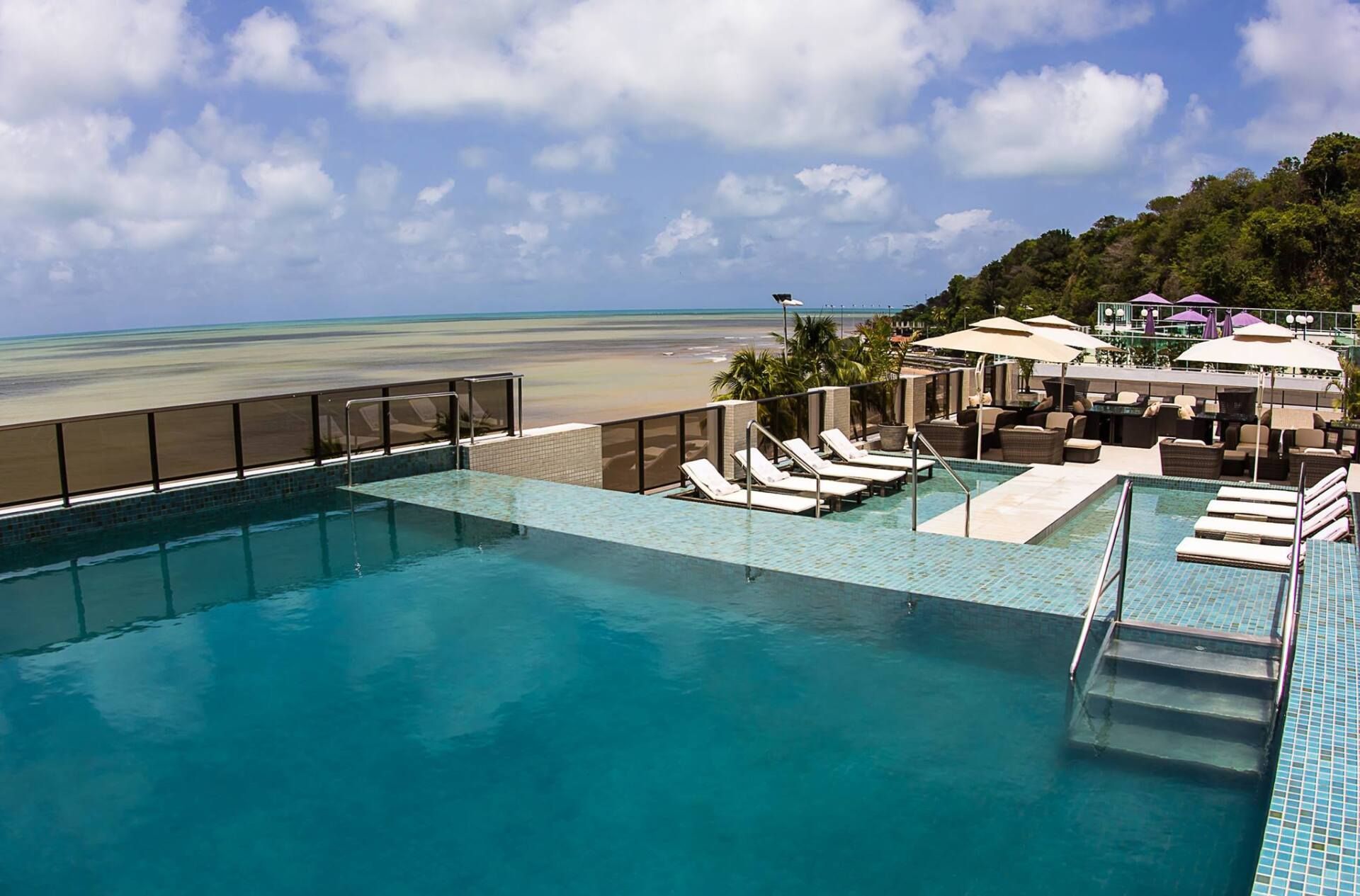 Piscina infinita com vista para o oceano, com espreguiçadeiras e edifícios num dia ensolarado.
