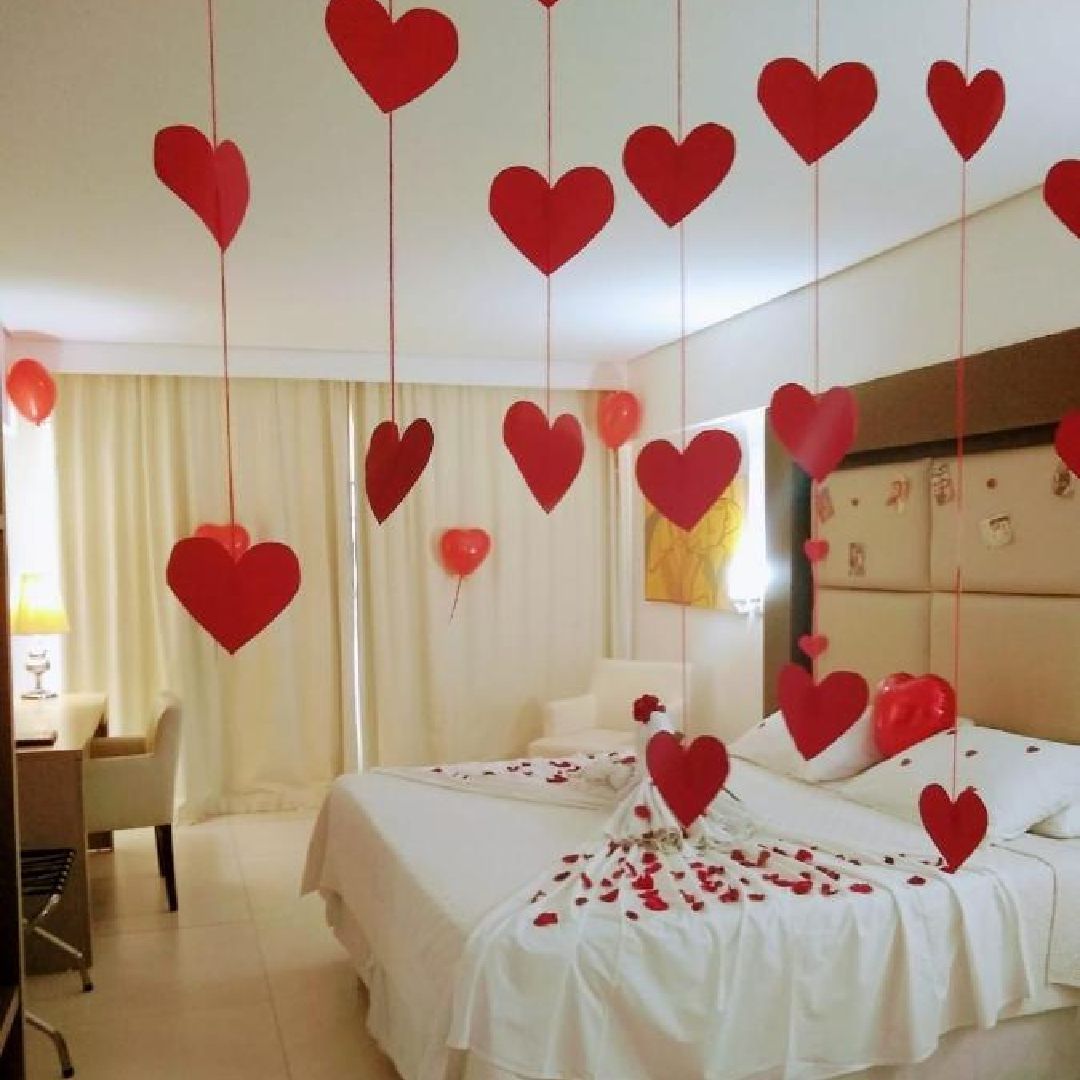Quarto decorado com corações de papel vermelho pendurados e pétalas de rosa na cama para uma ocasião romântica.