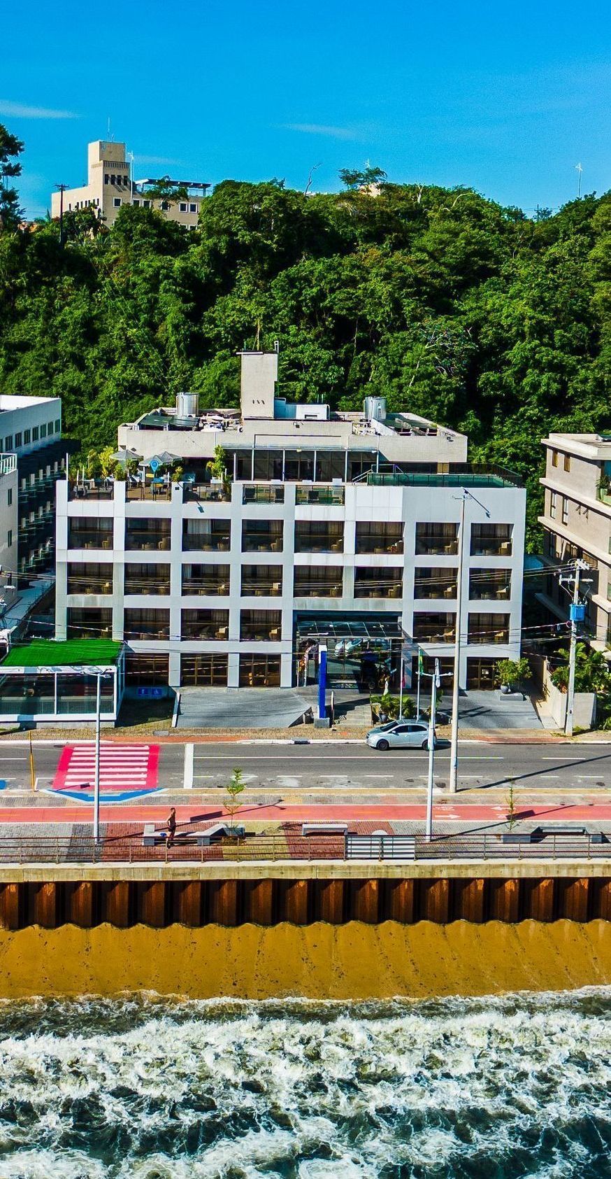 Prédio branco com muitas janelas perto de uma praia, com vegetação e céu azul ao fundo.