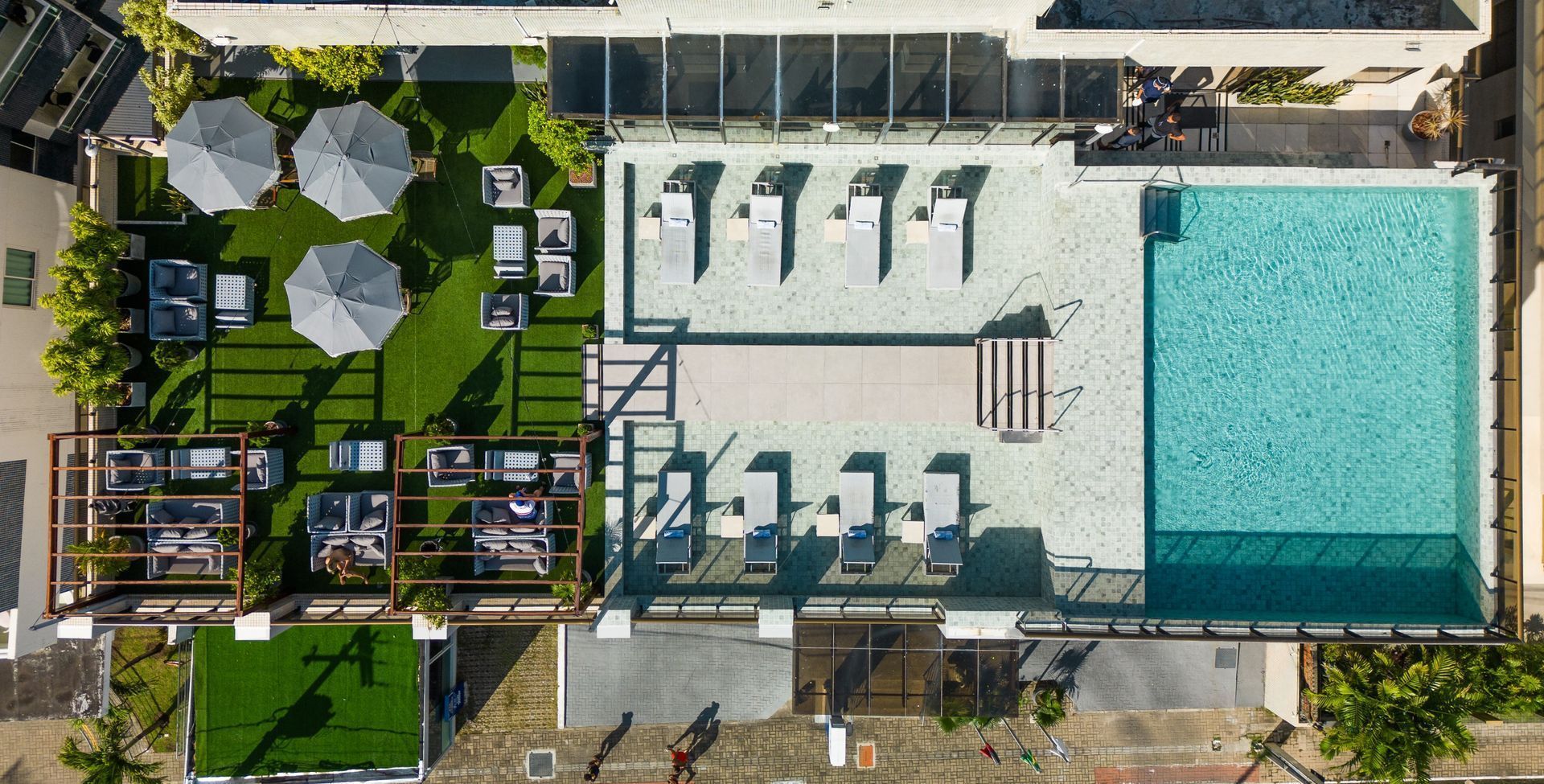 Vista aérea da área da piscina na cobertura, com espreguiçadeiras, guarda-sóis e mesas de jantar sobre grama artificial.