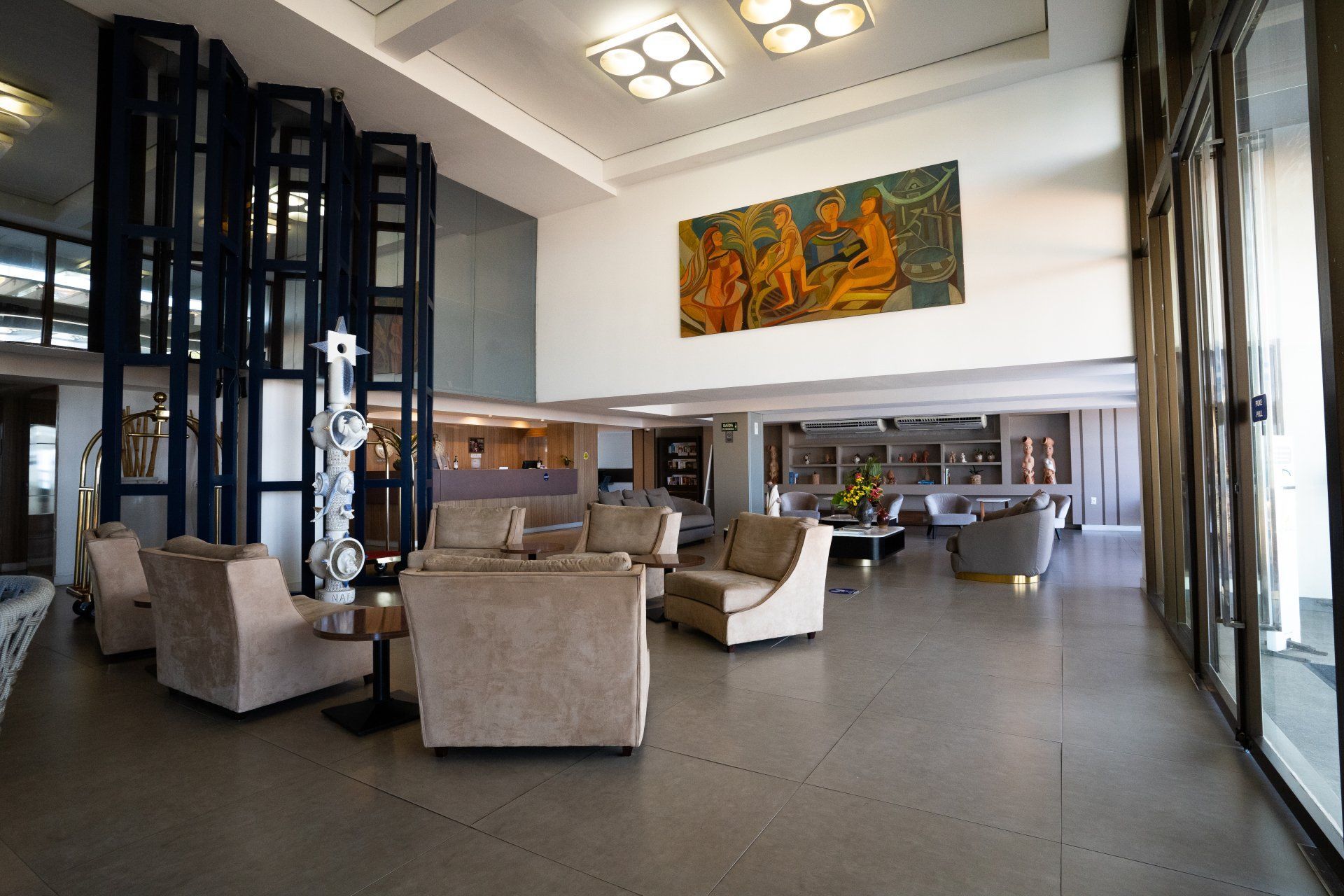 Hall de entrada com assentos bege, grandes obras de arte e uma estrutura de elevador estilizada em azul. Bem iluminado por grandes janelas.