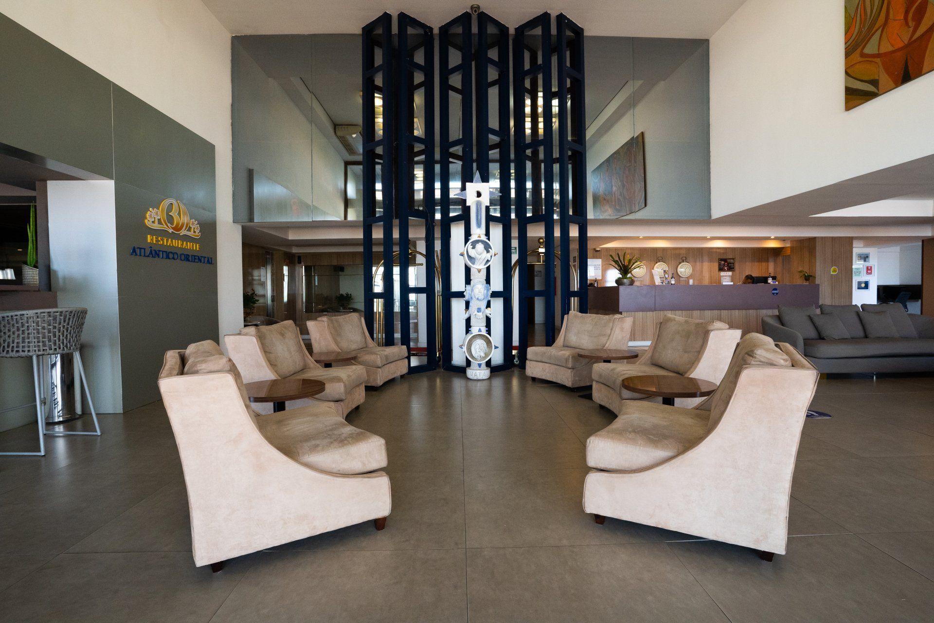 Hall de entrada com assentos ao redor de uma estrutura central escura com treliças. Sofás e mesas laterais em tons claros, ambiente luminoso.