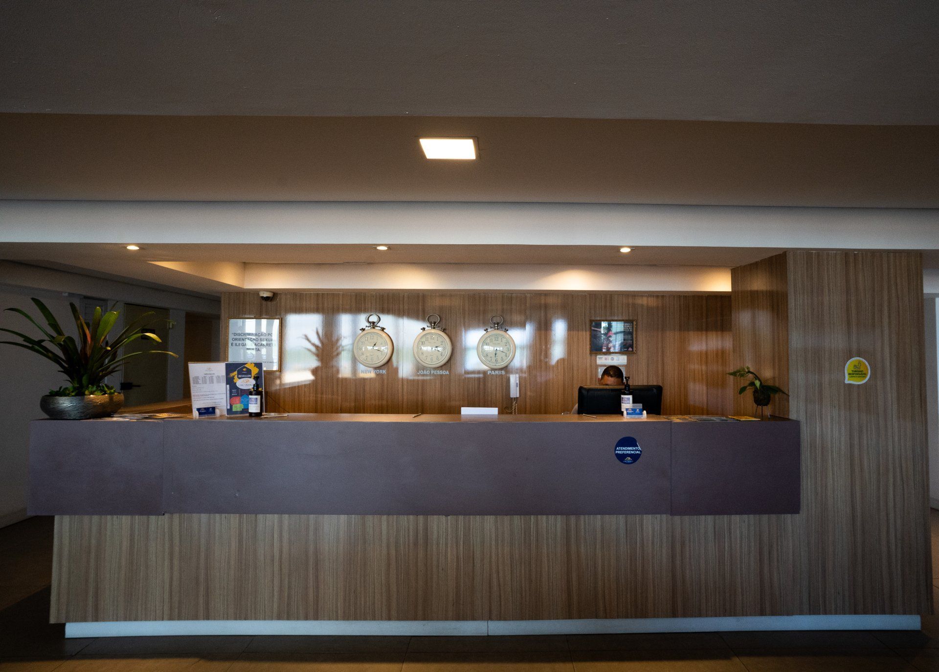 Recepção de hotel com relógios, pessoa trabalhando e painéis de madeira.