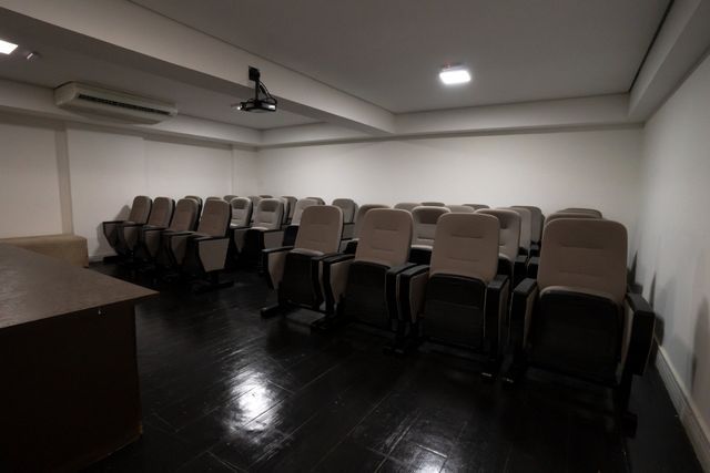 Auditório vazio com fileiras de cadeiras bege, piso preto, paredes brancas e um projetor.