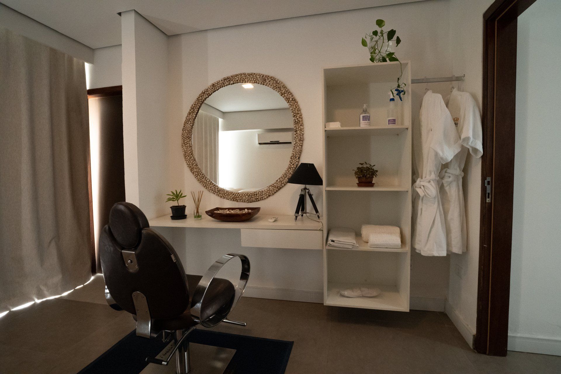 Interior de salão de beleza com cadeira, espelho, prateleira com produtos e roupões.