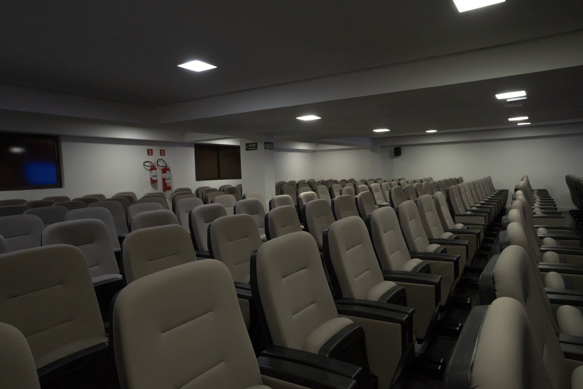 Fileiras de poltronas bege em um auditório vazio, iluminação suspensa, paredes e teto escuros.
