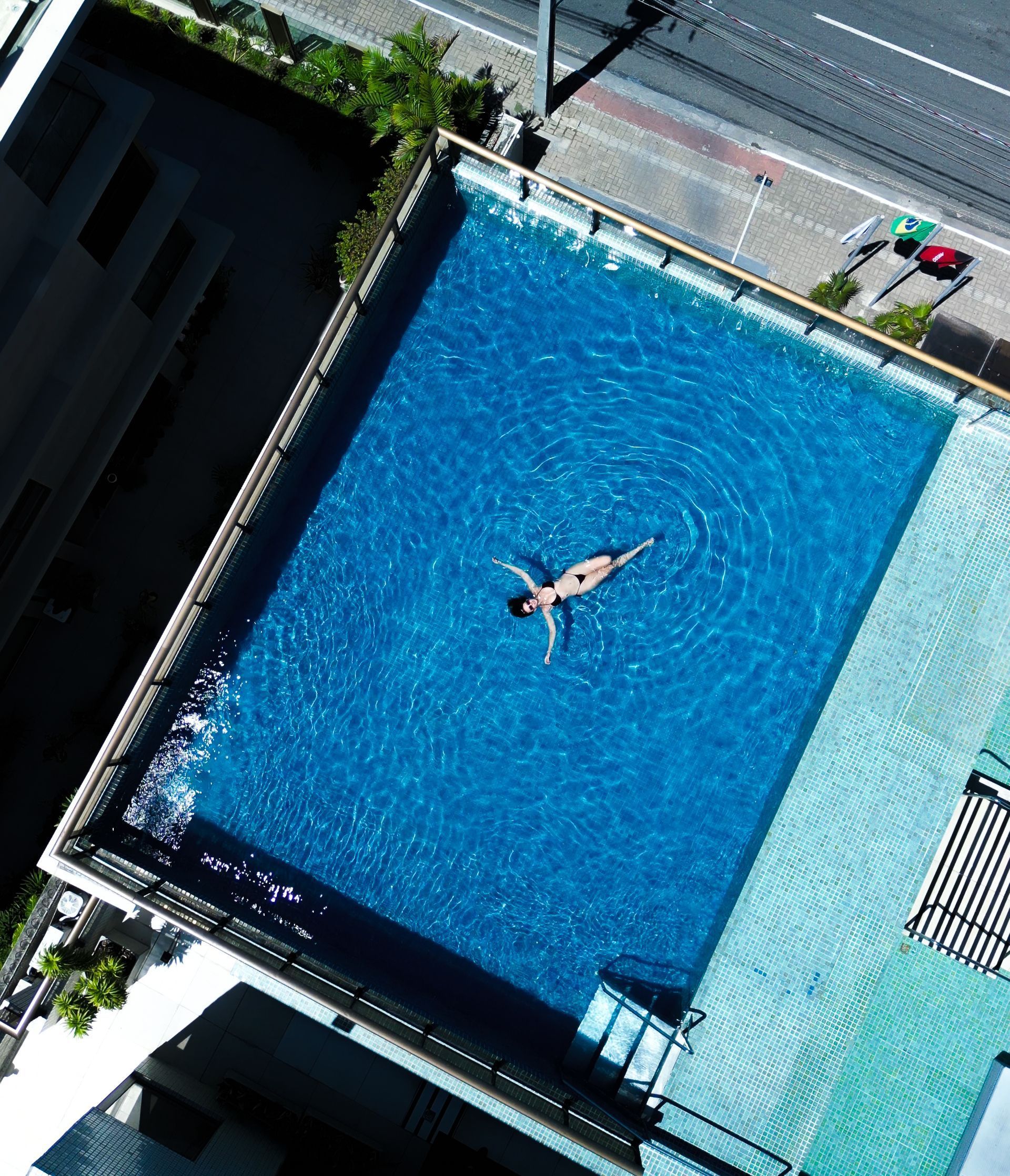 Piscina na cobertura com uma pessoa nadando. Água azul, prédios e a rua são visíveis de cima.