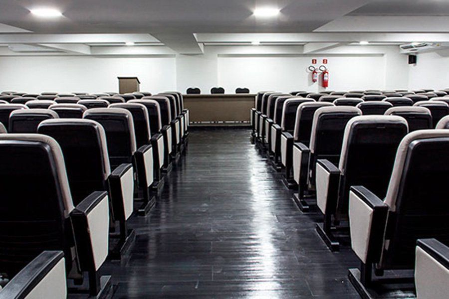 Fileiras de cadeiras pretas vazias em um auditório grande e iluminado. Um pequeno palco na frente.