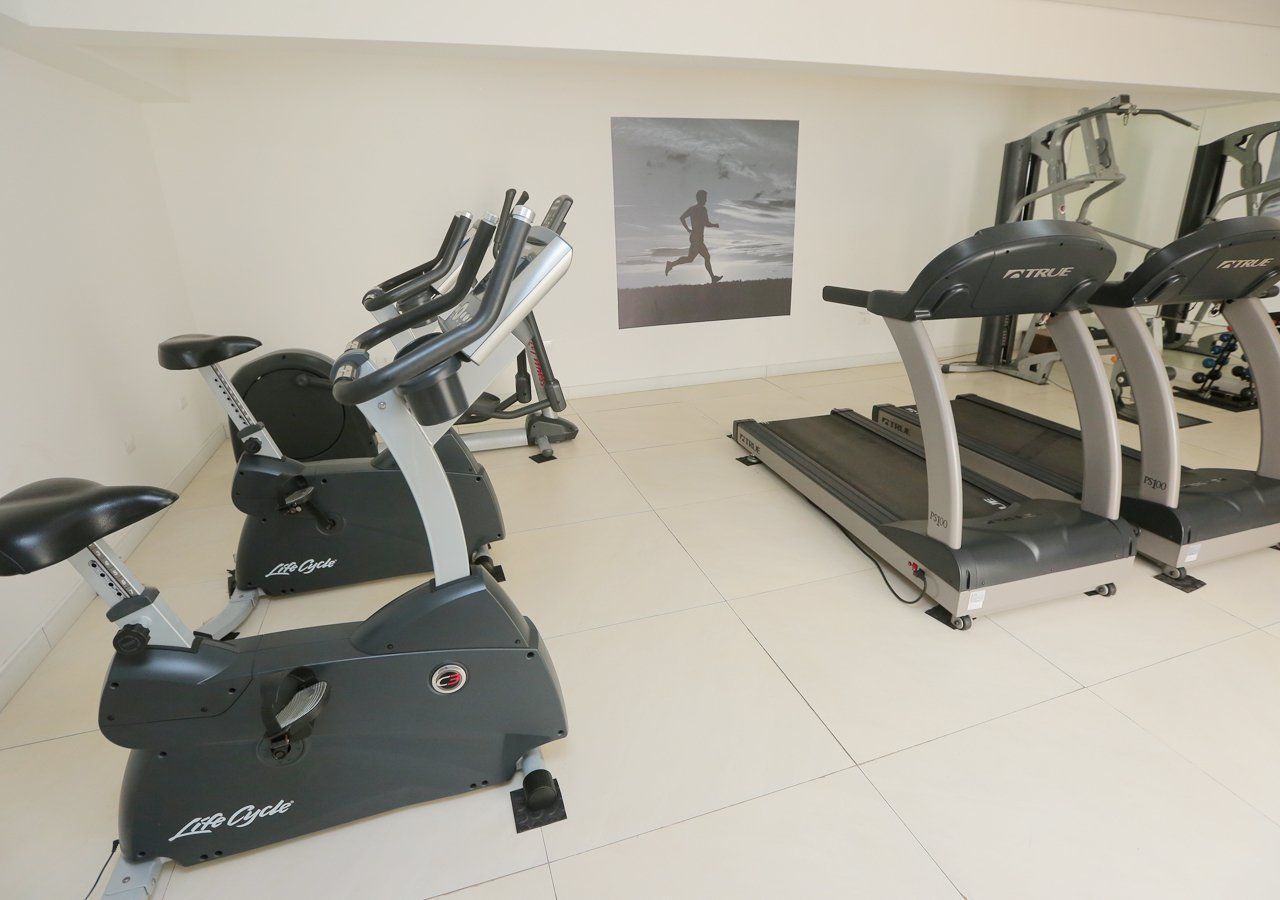 Interior de academia com bicicletas ergométricas, esteiras, aparelhos de musculação e um pôster sobre corrida.