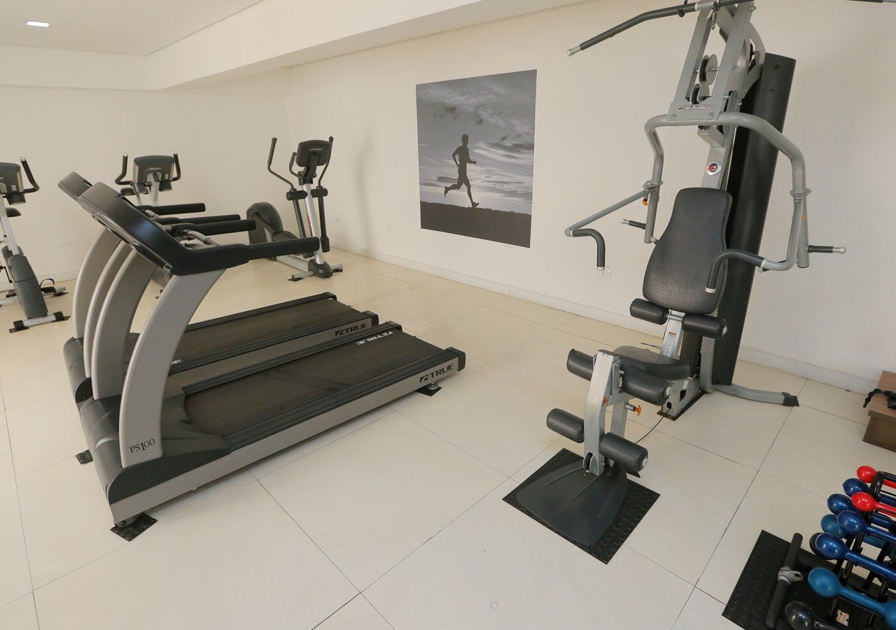 Interior de academia com esteira, aparelhos de ginástica, pesos e uma foto na parede.